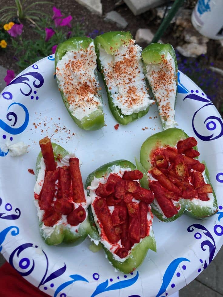 Vista superior de um prato de papel estampado com arabescos azuis contendo seis pedaços de pimentão verde recheados. Na parte superior, três pedaços estão apenas com cream cheese e tempero em pó, e na parte inferior, três pedaços estão recheados com cream cheese e cobertos generosamente com snacks Takis Fuego crocantes.
