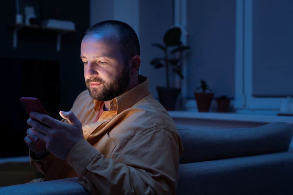 Homem jovem com capacete de proteção laranja e camisa xadrez, segurando um projeto de engenharia em papel em um canteiro de obras ensolarado.