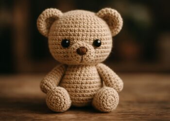 Urso amigurumi de crochê bege sentado sobre superfície de madeira com fundo escuro desfocado
