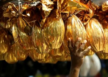 Mão tocando ovos de Páscoa dourados em embalagem metálica expostos em banca — bancos fecham no feriado de Páscoa sexta-feira 3 de abril 2026