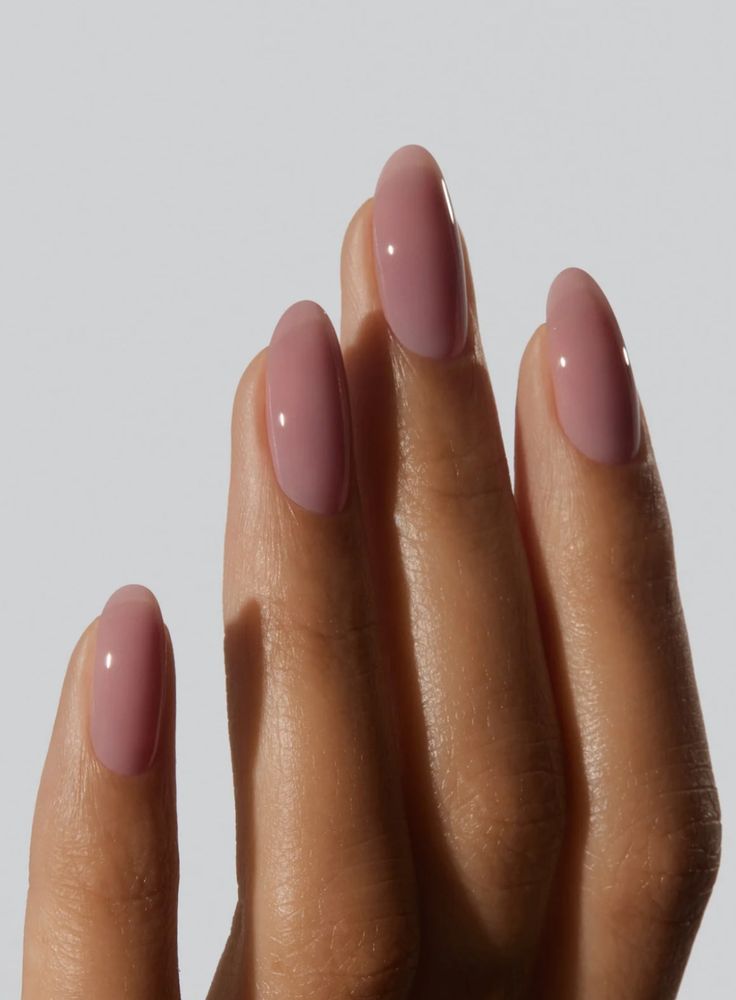 Close focado nos dedos de uma mão com pele morena, exibindo unhas longas e amendoadas pintadas com um esmalte bio-nude rosado translúcido, com um acabamento brilhante e gelatinoso (efeito jelly) sobre um fundo cinza claro uniforme.