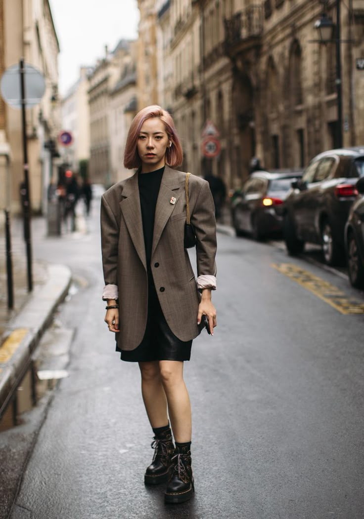 Mulher com cabelo rosa em look urbano com blazer oversized marrom de risca de giz, vestido preto curto e coturno preto tratorado.