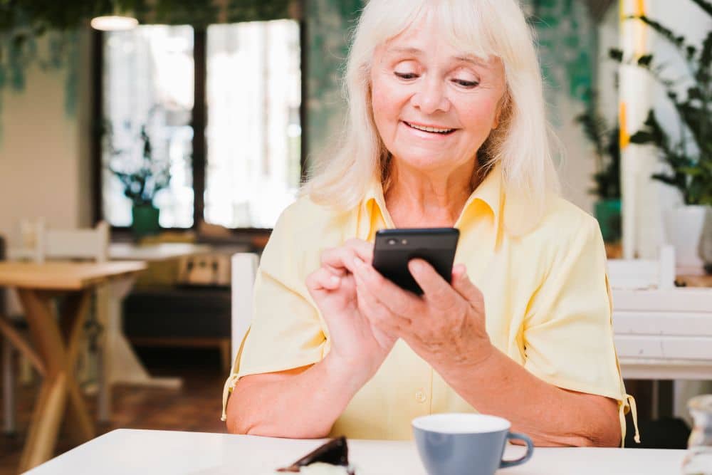 Mulher sênior sorridente com cabelos brancos, usando uma camisa amarela e sentada em uma mesa branca em um café. Ela segura um smartphone preto na vertical com as duas mãos e olha para a tela, com uma xícara de café azul em primeiro plano.