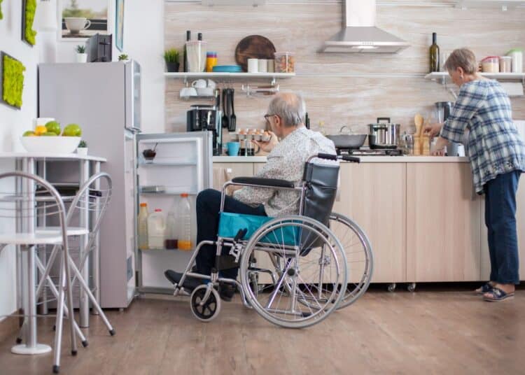 Fotografia de um homem idoso e grisalho sentado em uma cadeira de rodas azul, de costas para a câmera, com a porta da geladeira aberta à sua frente, tentando alcançar itens. Ao fundo, borrada, uma mulher idosa em pé trabalha em uma bancada de cozinha alta, evidenciando as barreiras arquitetônicas em um ambiente doméstico comum, ideal para ilustrar a necessidade de adaptações de acessibilidade.
