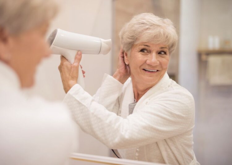 Mulher madura sorridente com cabelos curtos e grisalhos, vestindo um roupão branco, secando o cabelo com um secador branco em frente ao espelho do banheiro. A imagem ilustra a rotina de cuidados e secagem correta para evitar o amarelamento.