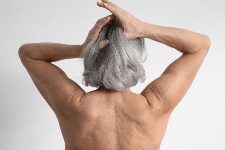 Close das costas e da nuca de uma mulher idosa com cabelos curtos e grisalhos. Suas mãos estão levantadas, tateando a textura do cabelo seco. O fundo é cinza claro e neutro. A imagem ilustra a aspereza causada pelo uso incorreto de produtos ou água quente.