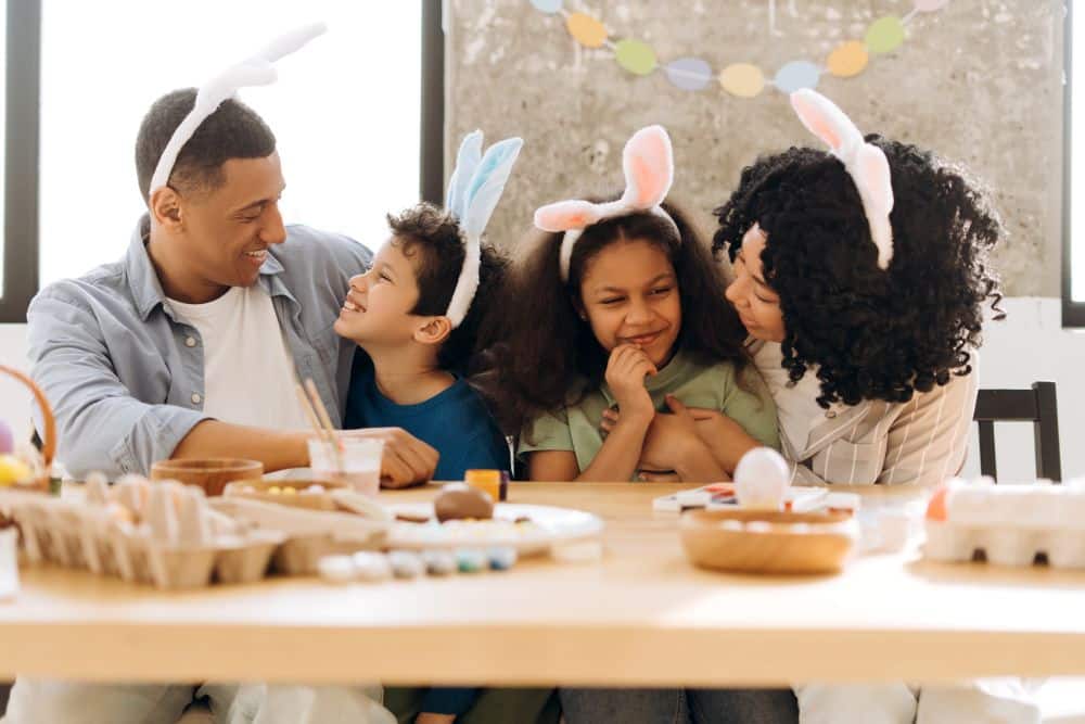 Close nas mãos de uma família, composta por pai de camisa jeans e mãe de cabelos cacheados, todos usando orelhas de coelho brancas, sentados ao redor de uma mesa de madeira. Eles estão sorrindo enquanto decoram ovos de Páscoa com confeitos e tintas, e no fundo, há um quadro com post-its e decorações de Páscoa.