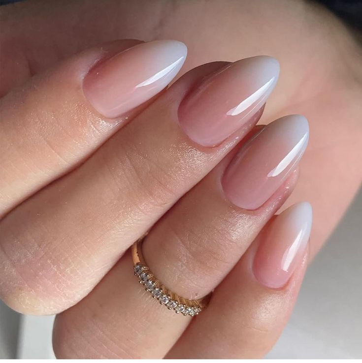 Close de unhas ovais com efeito francesinha difusa (baby boomer). O design apresenta um degradê suave entre a base nude rosada e as pontas em branco translúcido, com acabamento de alto brilho e um anel delicado no dedo anelar.