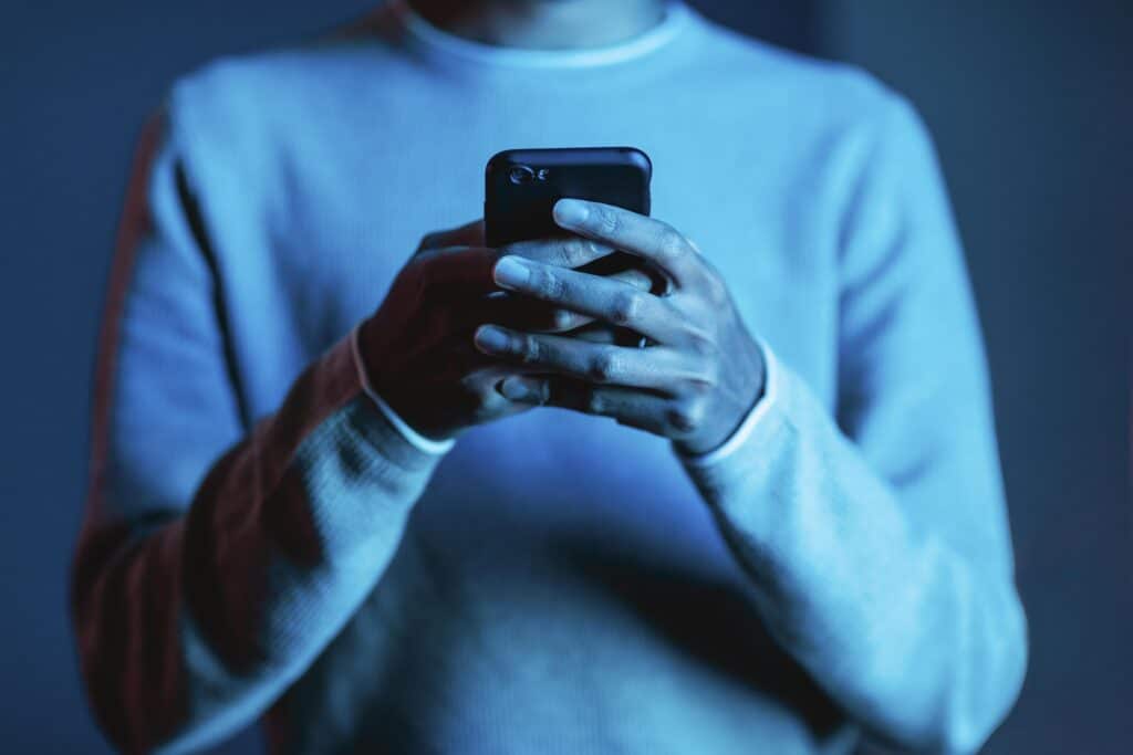 Close nas mãos de uma pessoa usando blusa azul, segurando um smartphone com as duas mãos e digitando, em um ambiente com iluminação cênica azul e vermelha.