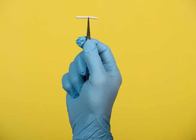 Close de uma mão usando luva cirúrgica azul e segurando uma pinça metálica que eleva um pequeno bastão branco de Implanon (implante contraceptivo subdérmico). O fundo é amarelo sólido e vibrante, destacando o dispositivo médico.