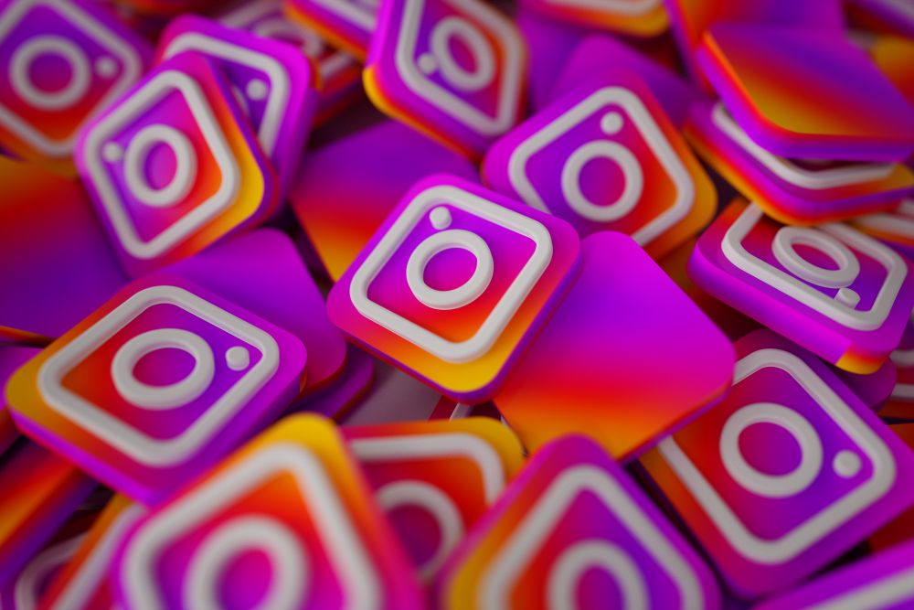 Diversos ícones tridimensionais do Instagram com as cores gradientes características (roxo, rosa e laranja) amontoados em um fundo infinito, criando um efeito de profundidade e repetição.