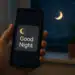 Mão segurando celular com mensagem 'Good Night' ao lado de uma janela à noite