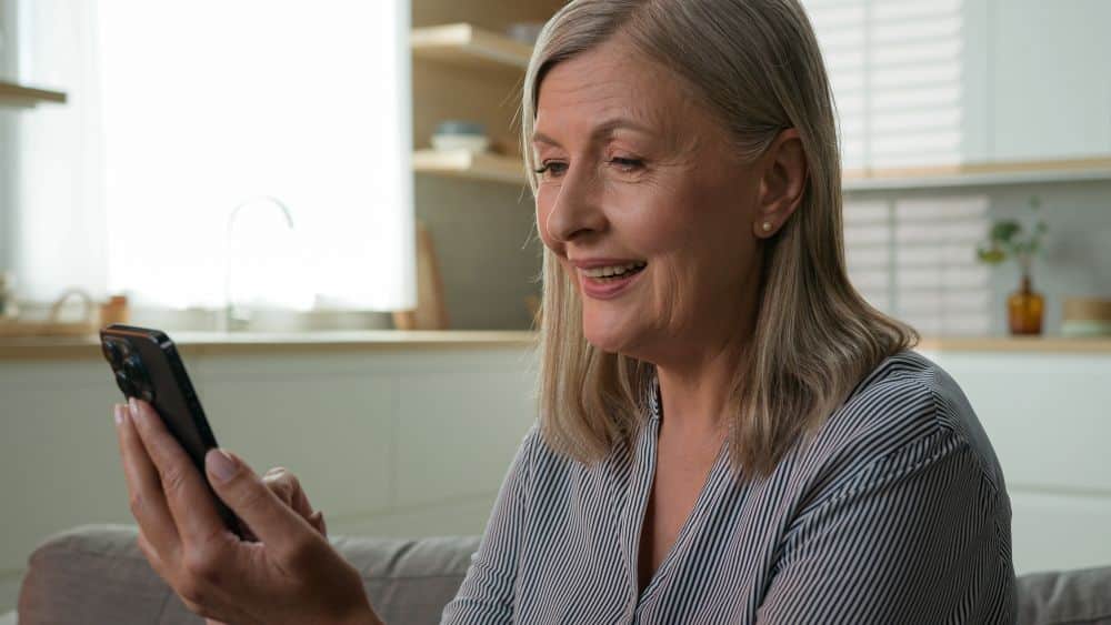 Close de uma mulher idosa sorridente, com cabelos loiros curtos, vestindo uma camisa listrada em tons de cinza e branco. Ela está segurando um smartphone preto com a mão esquerda e olhando para a tela com expressão de alegria, enquanto usa o polegar direito para navegar. O fundo é uma cozinha residencial clara e desfocada, com armários brancos e uma janela.