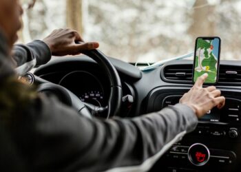 Foto interna de um motorista com o braço estendido tocando na tela de um smartphone que mostra um mapa de navegação em um suporte no painel do carro.
