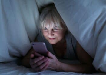 Mulher idosa de cabelos brancos sorrindo suavemente enquanto mexe em um smartphone rosa debaixo das cobertas brancas em um ambiente escuro à noite.