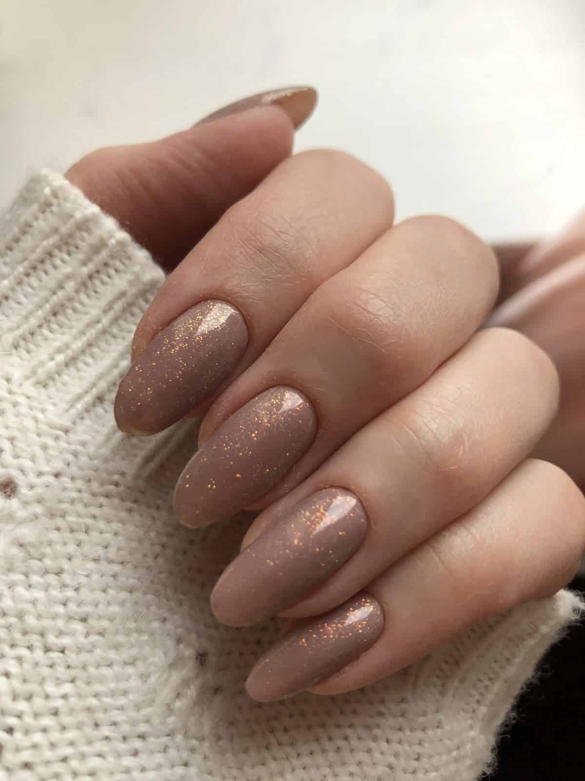 Mão feminina com unhas nude terroso e esmalte com brilho sutil apoiada sobre suéter claro