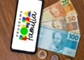 Um smartphone sobre uma mesa de madeira exibindo a logomarca colorida do Programa Bolsa Família na tela. Ao lado do celular, há notas de 20, 50 e 100 reais e algumas moedas espalhadas.
