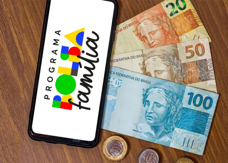 Um smartphone sobre uma mesa de madeira exibindo a logomarca colorida do Programa Bolsa Família na tela. Ao lado do celular, há notas de 20, 50 e 100 reais e algumas moedas espalhadas.