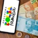 Um smartphone sobre uma mesa de madeira exibindo a logomarca colorida do Programa Bolsa Família na tela. Ao lado do celular, há notas de 20, 50 e 100 reais e algumas moedas espalhadas.