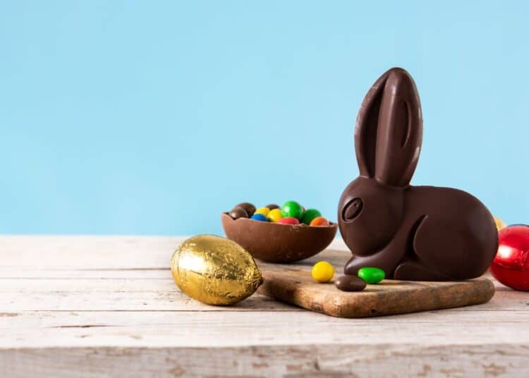 Close em um coelho de chocolate ao leite em pé em uma tábua de madeira rústica, ao lado de um ovo de chocolate aberto cheio de confeitos coloridos. À esquerda, um ovo de chocolate embalado em papel dourado brilha sobre a mesa de madeira clara. O fundo é um azul suave.