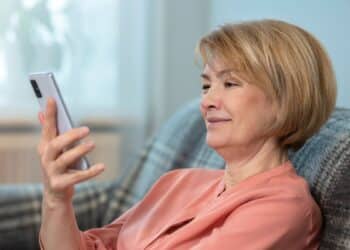 Mulher madura de cabelos loiros curtos, vestindo uma blusa salmão, sentada em um sofá cinza com manta xadrez. Ela segura um smartphone prateado com a mão esquerda e sorri suavemente enquanto olha para a tela, em um ambiente doméstico iluminado e acolhedor.