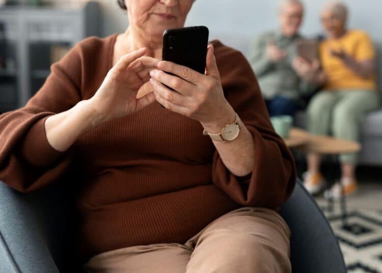 Close focado nas mãos de uma mulher idosa de blusa marrom e relógio, sentada em uma poltrona azul e segurando um smartphone preto na vertical. Ao fundo, desfocados, um homem e outra mulher mais velhos estão sentados em um sofá.