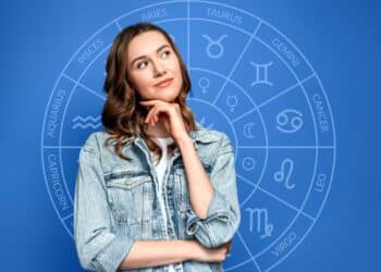 : Uma mulher jovem com expressão pensativa, tocando o queixo, à frente de um fundo azul com um gráfico circular dos signos do zodíaco e seus símbolos.