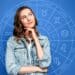 : Uma mulher jovem com expressão pensativa, tocando o queixo, à frente de um fundo azul com um gráfico circular dos signos do zodíaco e seus símbolos.