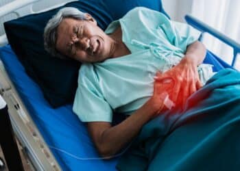 Homem idoso deitado em uma cama de hospital, expressando dor e segurando a região do abdômen com um brilho avermelhado indicando o local do desconforto.