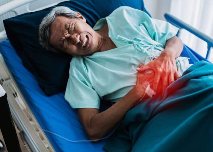 Homem idoso deitado em uma cama de hospital, expressando dor e segurando a região do abdômen com um brilho avermelhado indicando o local do desconforto.