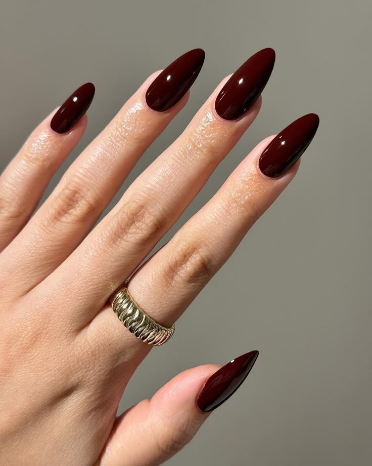 Mão feminina com unhas longas no formato stiletto pintadas com esmalte Burgundy (vinho escuro) de acabamento ultra brilhante, usando um anel largo dourado com textura de gomos no dedo anelar.