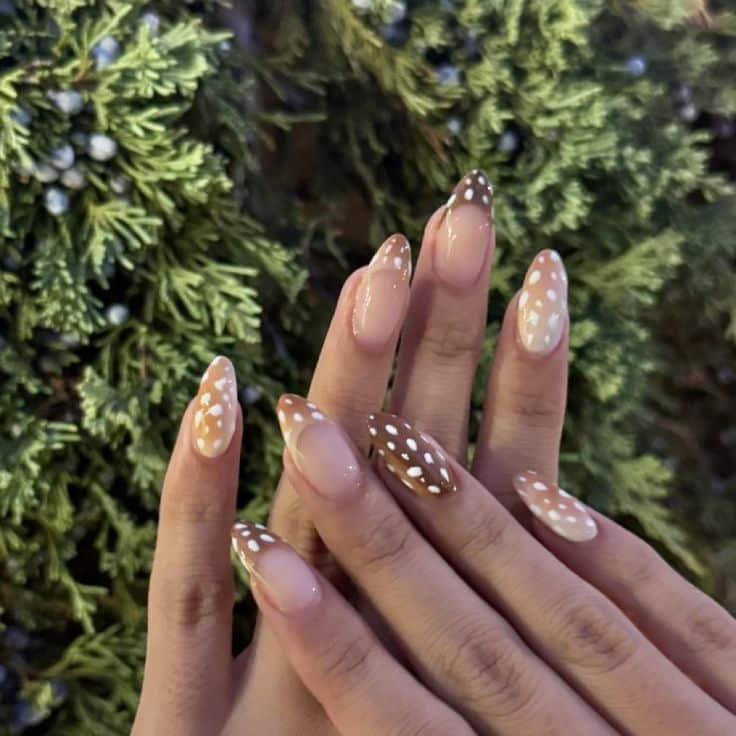 Mãos femininas com unhas amendoadas exibindo nail art de estampa de cervo. O design utiliza tons de marrom e bege com pintas brancas irregulares, alternando entre cobertura total e francesinhas largas, com fundo de folhagens verdes.