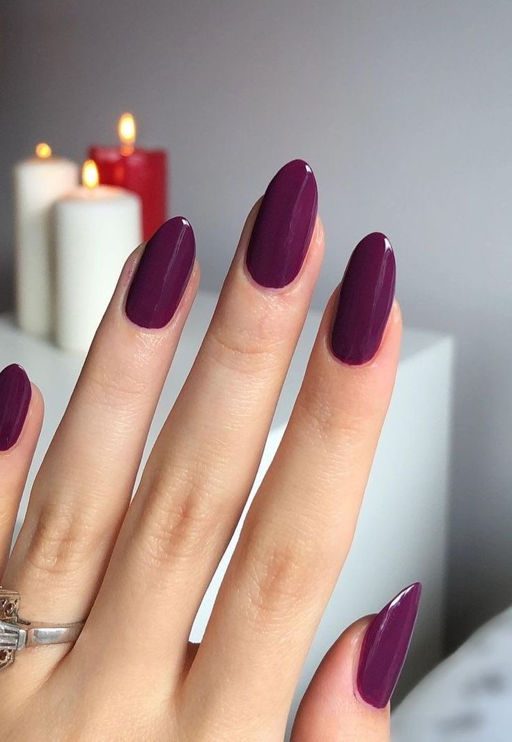 Mão feminina com unhas longas e amendoadas pintadas em esmalte roxo berinjela brilhante, com um anel de prata delicado no dedo anelar e velas acesas ao fundo desfocado.