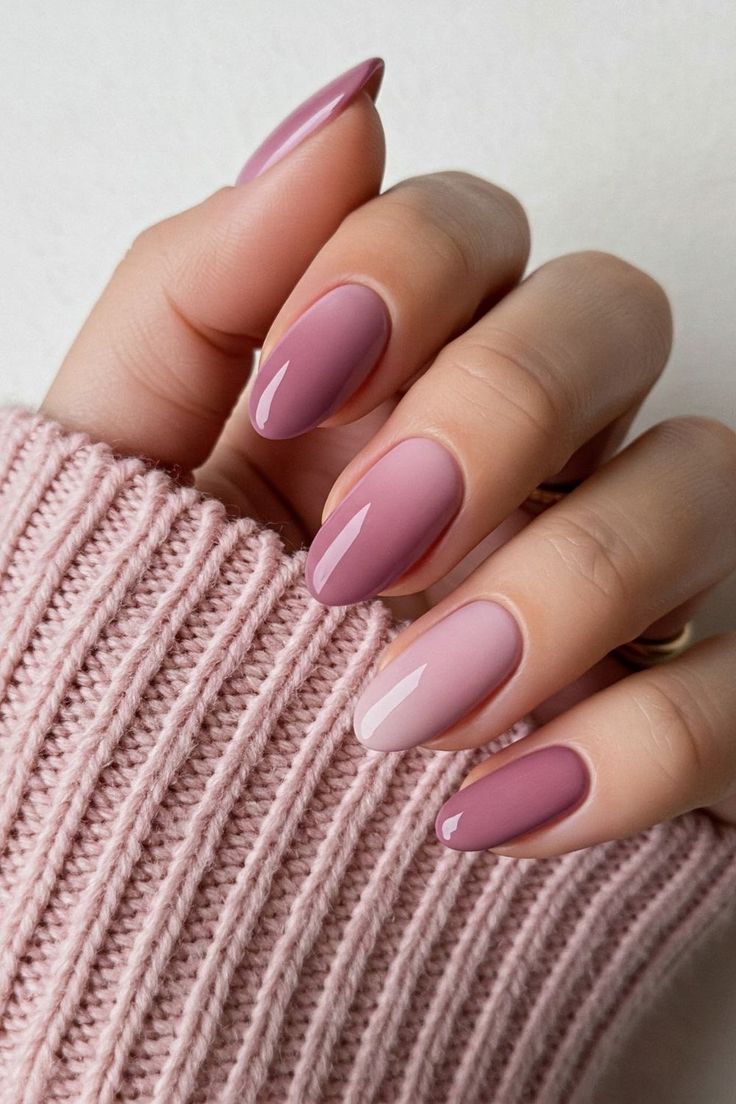 Mão feminina com unhas ovais pintadas em diferentes tonalidades de rosa e malva, criando um efeito degradê suave entre os dedos. A mão repousa sobre a manga de um tricô rosa claro.