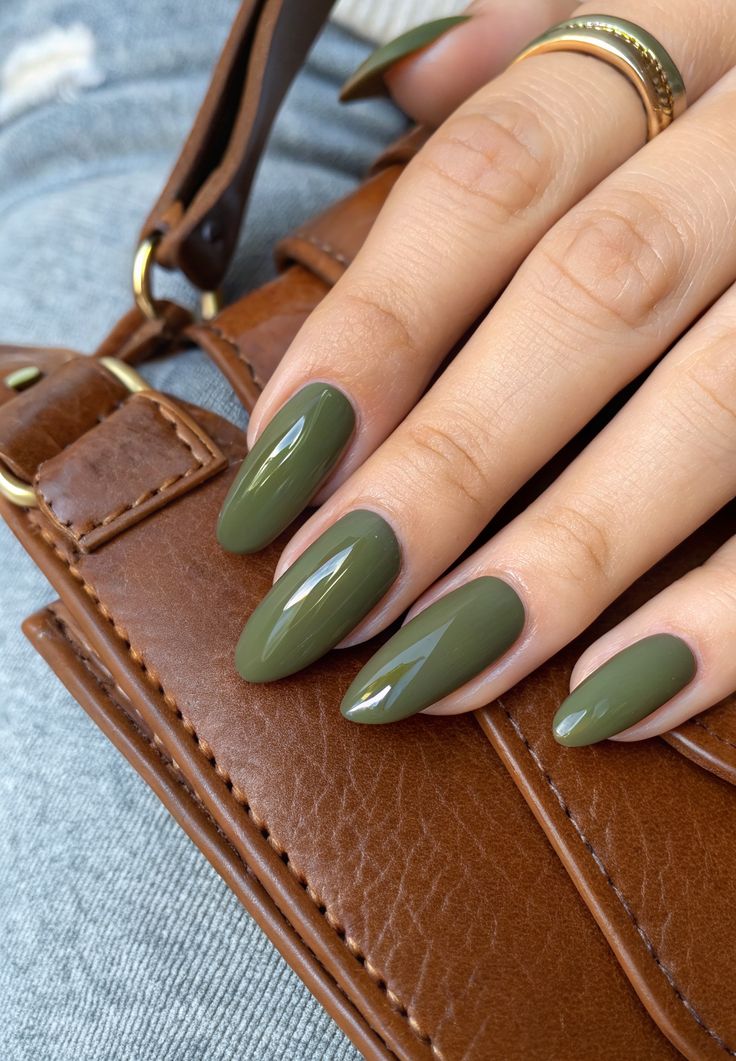 Close de uma mão feminina com unhas longas amendoadas pintadas em esmalte verde oliva brilhante, apoiada sobre uma bolsa de couro marrom, usando um anel dourado.