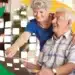 Um casal de idosos sorridentes, um homem sentado e uma mulher de pé ao seu lado, olhando para a tela de um notebook. A imagem possui elementos gráficos em verde e amarelo e pequenos quadrados brancos que sugerem conectividade digital.