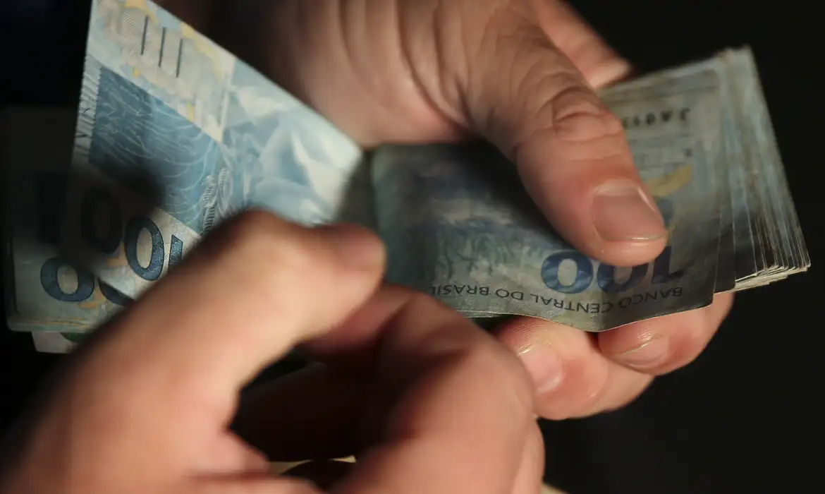 Mãos de uma pessoa contando diversas cédulas de cem reais brasileiros, representando o pagamento de benefícios do INSS e BPC.