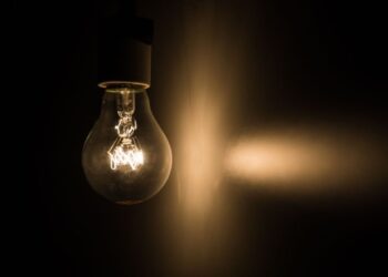 Uma lâmpada incandescente acesa em um ambiente escuro, emitindo uma luz amarelada que projeta um feixe lateral.