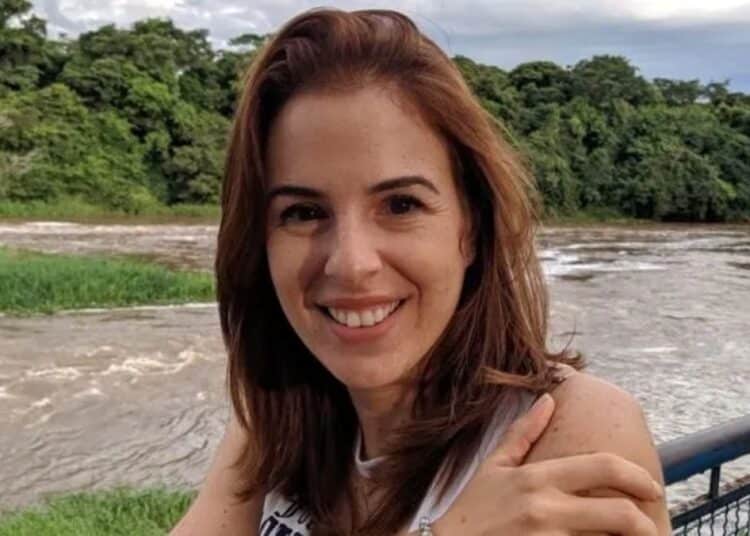 Suzane von Richthofen, com cabelos ruivos, sorrindo e olhando para a câmera. Ela está ao ar livre, com o braço dobrado, apoiada em uma grade azul. Ao fundo, vê-se um rio de águas turvas e uma densa vegetação verde sob céu nublado.
