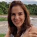 Suzane von Richthofen, com cabelos ruivos, sorrindo e olhando para a câmera. Ela está ao ar livre, com o braço dobrado, apoiada em uma grade azul. Ao fundo, vê-se um rio de águas turvas e uma densa vegetação verde sob céu nublado.