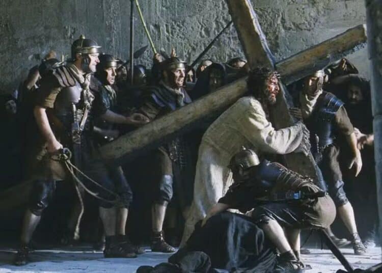 Cena cinematográfica da Via Crucis com Jesus carregando a cruz de madeira no ombro, cercado por soldados romanos em armaduras brônzeas e multidão ao fundo.