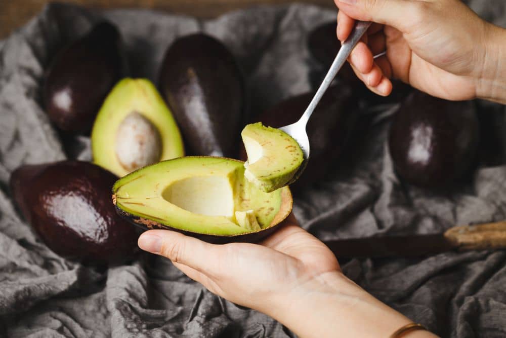 Close das mãos de uma pessoa retirando uma fatia generosa de um abacate cortado ao meio com uma colher de metal. Ao fundo, vários abacates inteiros com casca escura estão dispostos sobre um tecido cinza rústico. A polpa da fruta está madura, apresentando um tom verde vibrante e textura cremosa.