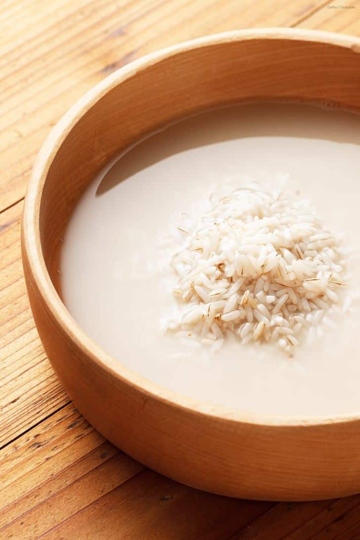 Recipiente de madeira contendo grãos de arroz cru submersos em água esbranquiçada e leitosa, demonstrando o processo de lavagem para extrair o amido.