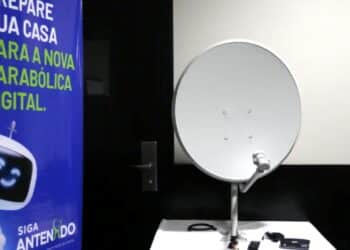 Uma antena parabólica digital branca montada sobre uma mesa, ao lado de um banner azul com a frase 'Prepare sua casa para a nova parabólica digital' e o logotipo Siga Antenado.