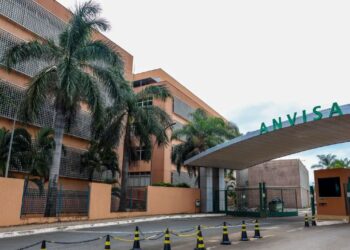 Vista externa da entrada principal da sede da ANVISA (Agência Nacional de Vigilância Sanitária). A imagem mostra o portal de acesso com o letreiro verde grande "ANVISA" e o edifício administrativo ao fundo, com palmeiras e grades de proteção.