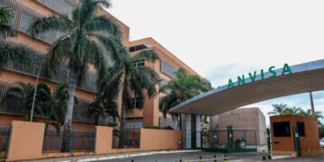 Vista externa da entrada principal da sede da ANVISA (Agência Nacional de Vigilância Sanitária). A imagem mostra o portal de acesso com o letreiro verde grande "ANVISA" e o edifício administrativo ao fundo, com palmeiras e grades de proteção.
