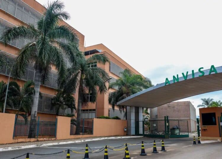 Vista externa da entrada principal da sede da ANVISA (Agência Nacional de Vigilância Sanitária). A imagem mostra o portal de acesso com o letreiro verde grande "ANVISA" e o edifício administrativo ao fundo, com palmeiras e grades de proteção.