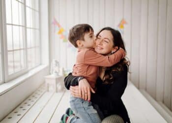 Uma mulher sorridente sentada no chão de uma sala clara, abraçando um menino pequeno que lhe dá um beijo na bochecha. Ao fundo, uma janela grande e bandeirinhas coloridas decoram a parede branca.