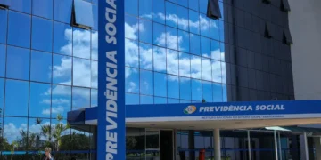 Fachada do edifício-sede do Instituto Nacional do Seguro Social (INSS) com vidros espelhados azuis refletindo o céu. Em primeiro plano, uma pilastra azul com a inscrição vertical "Previdência Social" e a placa oficial na entrada.