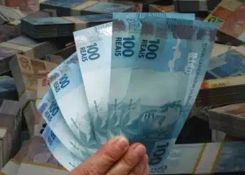 Pessoa segurando notas de cem reais em frente a dinheiro